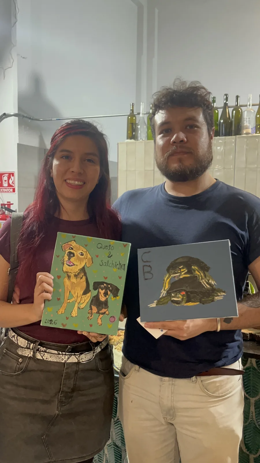 Una pareja muestra dos retratos de mascotas terminados en un evento de pinta y brinda