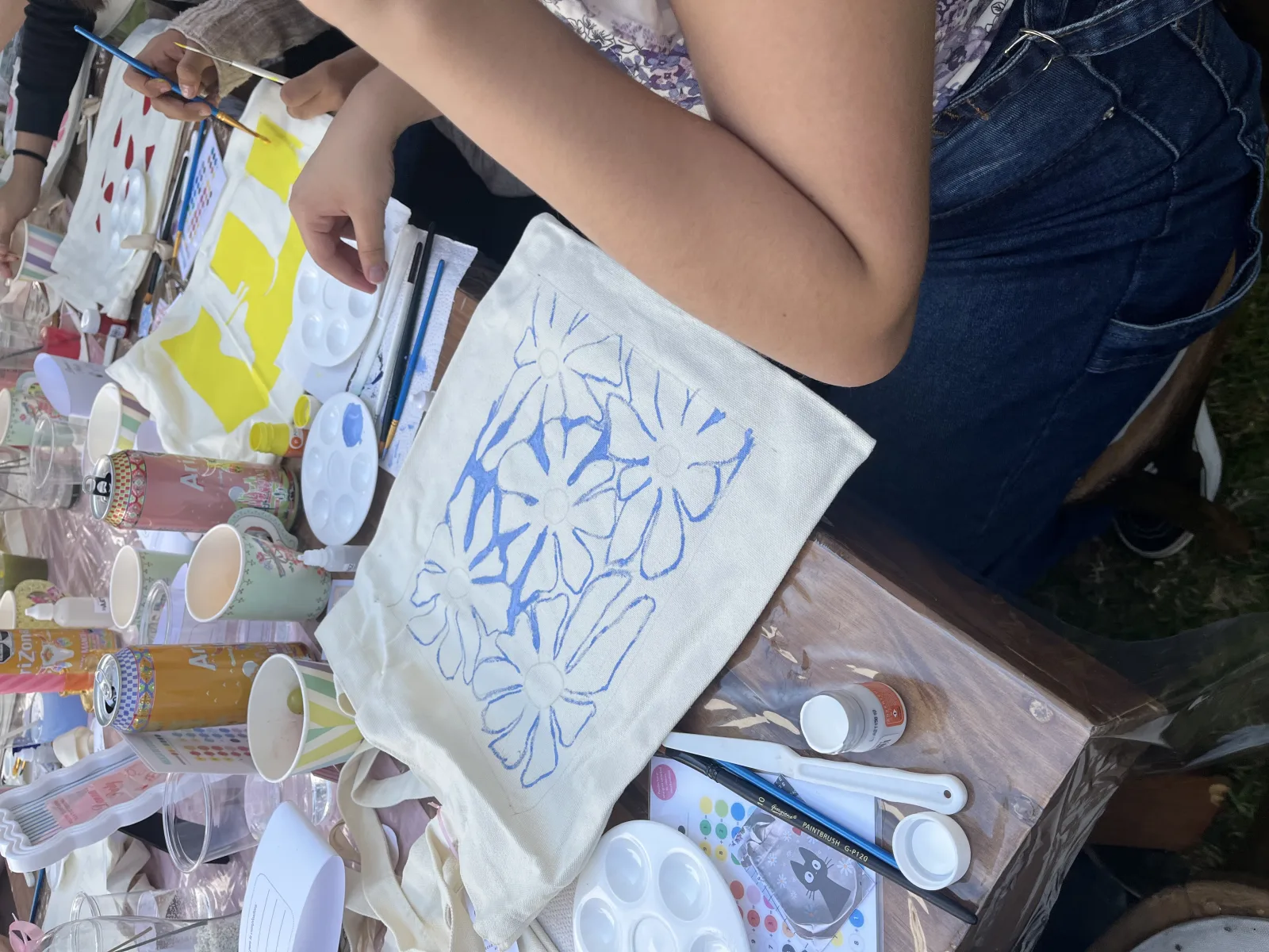 Una asistente pinta flores sobre una tote bag de tela en un taller al aire libre